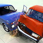 Zderzenie niebieskiego i czerwonego samochodu z widocznymi iskrami oraz stłuczonymi szybami