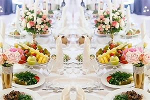 Elegancko nakryty stół z białymi obrusami, udekorowany świeżymi kwiatami, świecami i talerzami z owocami oraz warzywami