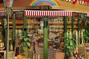 Kawiarnia z kolorowym tęczowym napisem, czerwono-białą markizą, ozdobnymi roślinami i stolikami na zewnątrz