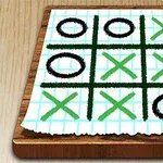 Kwadratowa kartka papieru z narysowaną siatką do gry, z zielonymi i czarnymi symbolami X i O, leżąca na drewnianej powierzchni